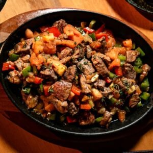 Delicious grilled beef with peppers and tomatoes in a hot skillet on wooden table.
